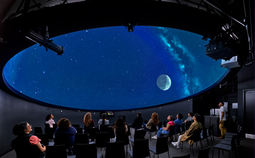 Students sitting in chairs looking at the the moon and stars on a screen in the planetarium